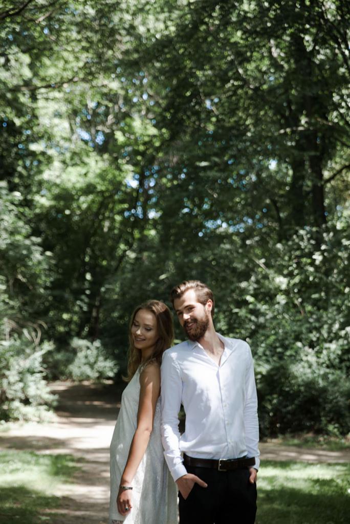 Uśmiechnięta para, oparta plecami o siebie, w lesie; kobieta w białej sukience, mężczyzna w białej koszuli i czarnych spodniach.