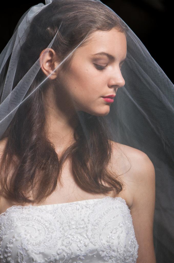 Portret panny młodej z profilu, z welonem na twarzy, w białej sukni z koronkowymi zdobieniami, na ciemnym tle.