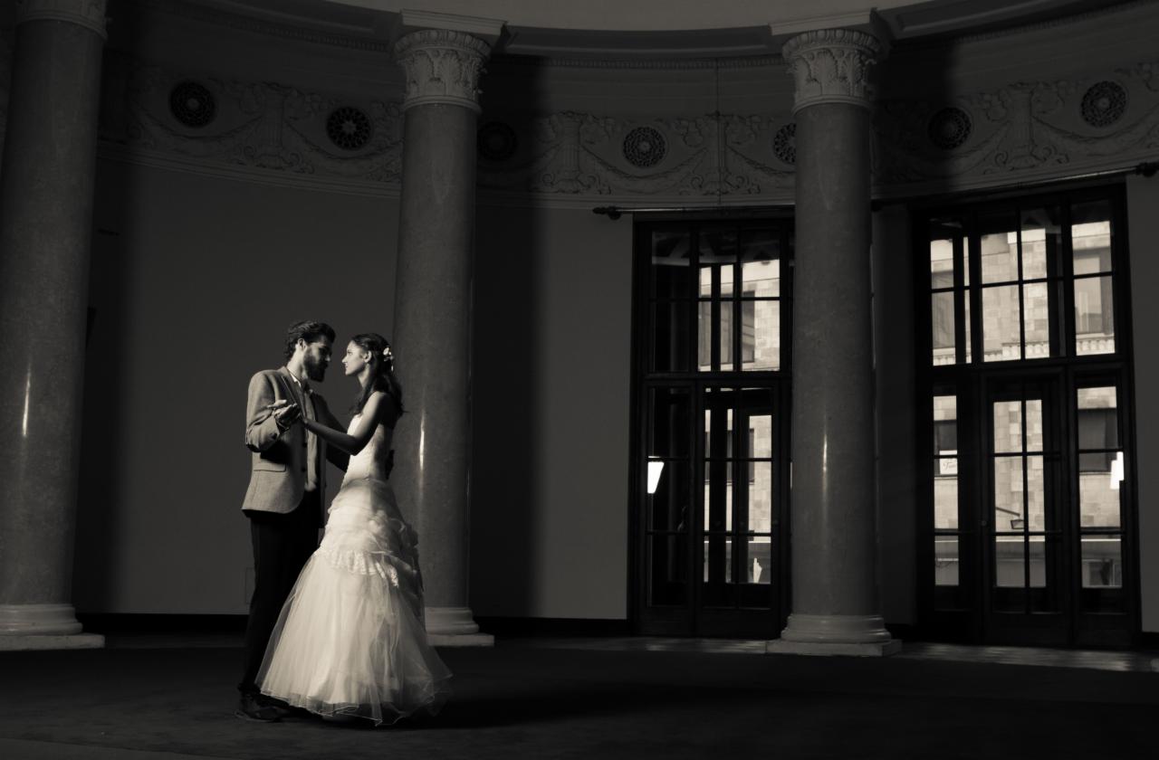 Czarno-białe zdjęcie pary młodej tańczącej w przestronnym holu z kolumnami i wysokimi oknami, suknia panny młodej z falbanami, pan młody w tweedowej marynarce.