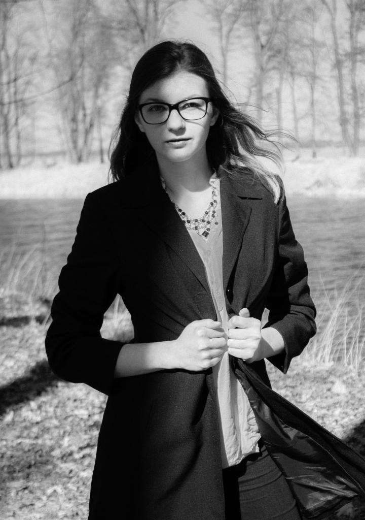 Portret młodej kobiety w okularach, ubrana w płaszcz, stojącej na tle rzeki i drzew, w czerni i bieli.