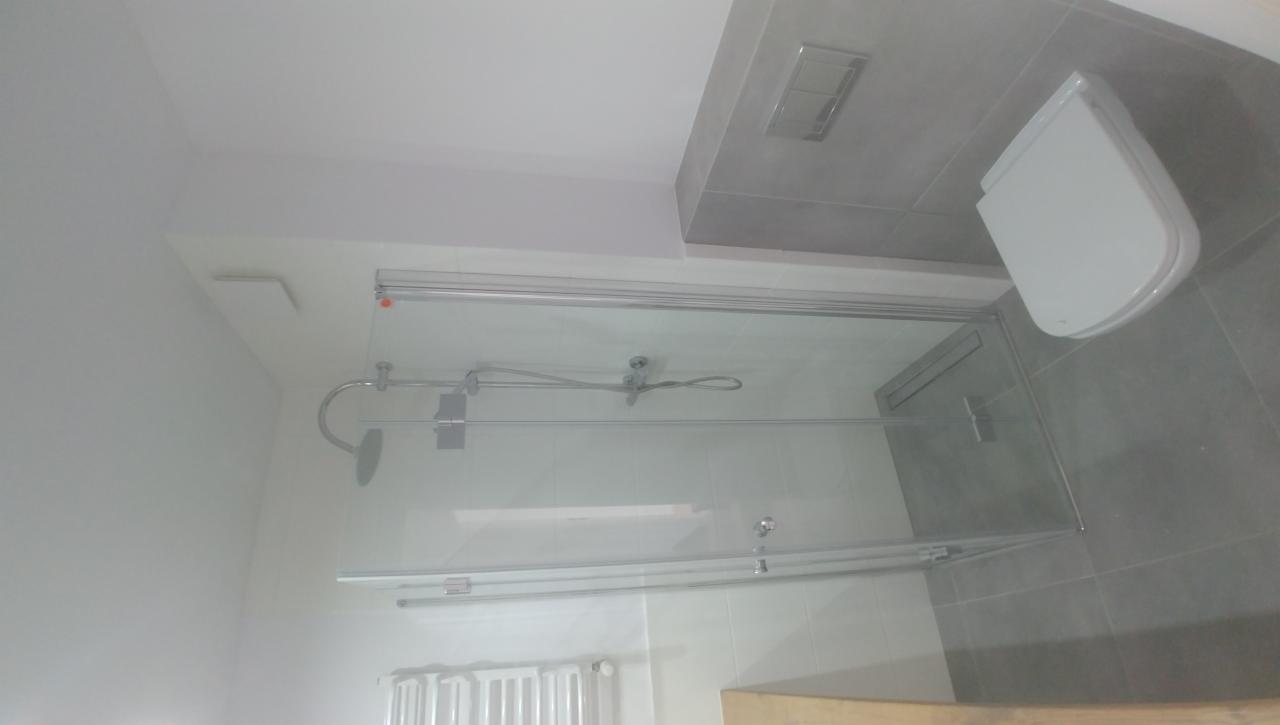 Nowoczesna kabina prysznicowa z przesuwnymi drzwiami, widok z góry na wykończoną łazienkę z szarymi płytkami i podwieszaną toaletą.