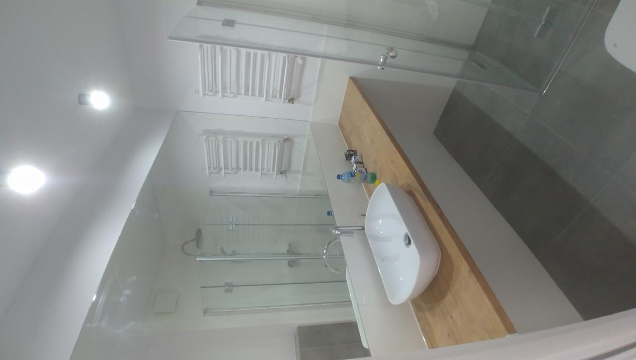 Nowoczesna łazienka z przeszkloną kabiną prysznicową, białą umywalką nablatową na drewnianym blacie i grzejnikiem drabinkowym na ścianie.