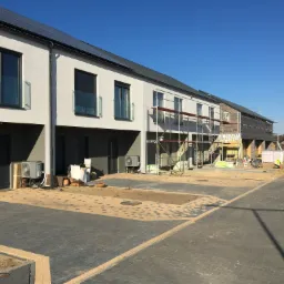 Rząd nowoczesnych domów szkieletowych w trakcie budowy, widoczne rusztowania, ocieplenie elewacji i panele słoneczne na dachu.