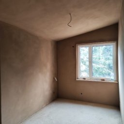 BUD-SOW - Pokój po nałożeniu gładzi, widoczne ściany i sufit w kolorze beżowym, okno z widokiem na zieleń. Wystający kabel na suficie.