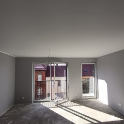 BUD-SOW - Pomalowane na szaro wnętrze pokoju z dużymi oknami balkonowymi i surową podłogą. Widoczne promienie słoneczne wpadające do środka. Świeżo wykończone ściany.