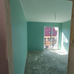 BUD-SOW - Pomieszczenie po malowaniu ścian na turkusowo, widoczne surowe wykończenie podłogi i ościeżnicy. W tle okno balkonowe z widokiem na budynek.