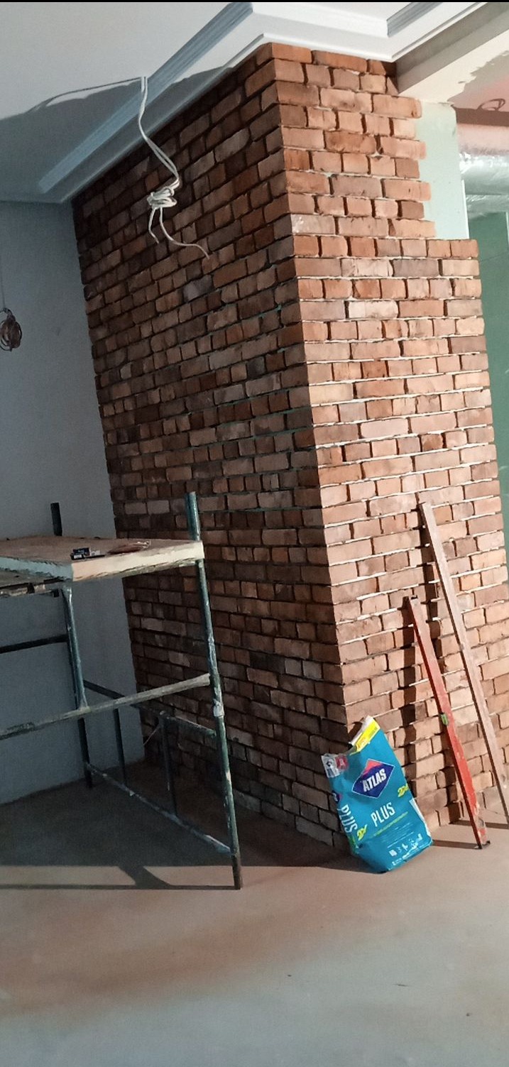 Wnętrze w trakcie prac wykończeniowych: ściana wyłożona cegłą klinkierową, widoczne rusztowanie, worki zaprawy, nieukończone prace przy suficie.