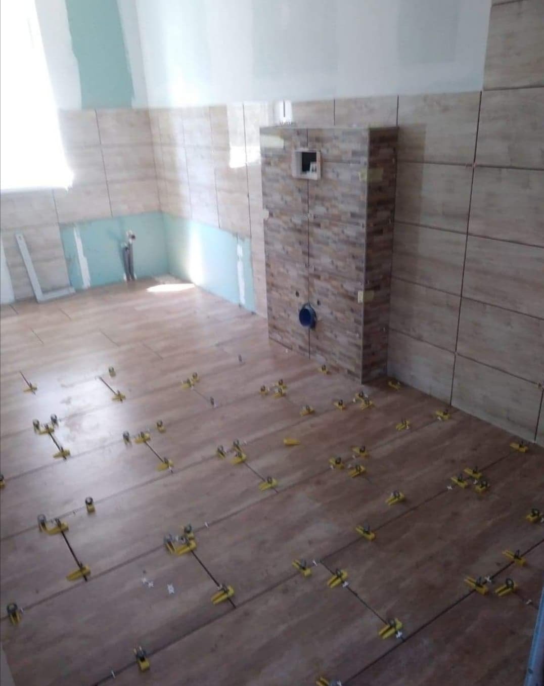 Wnętrze łazienki w trakcie wykańczania, z beżowymi płytkami na ścianach i podłodze, system poziomowania płytek widoczny na podłodze, fragment ściany z instalacją sanitarną w tle.