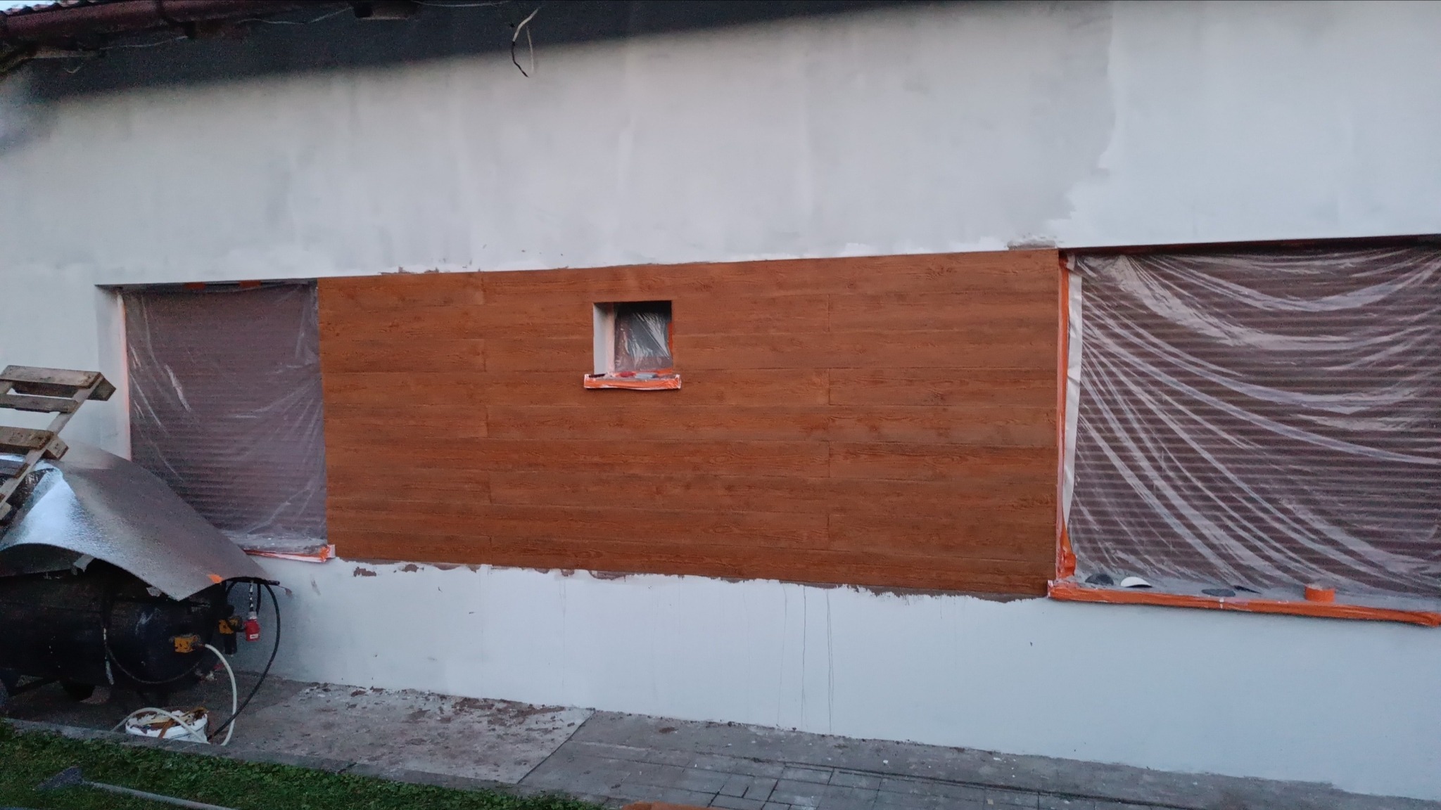 Fragment elewacji budynku z drewnopodobną okładziną, okna zakryte folią ochronną, widoczny kompresor i palety.