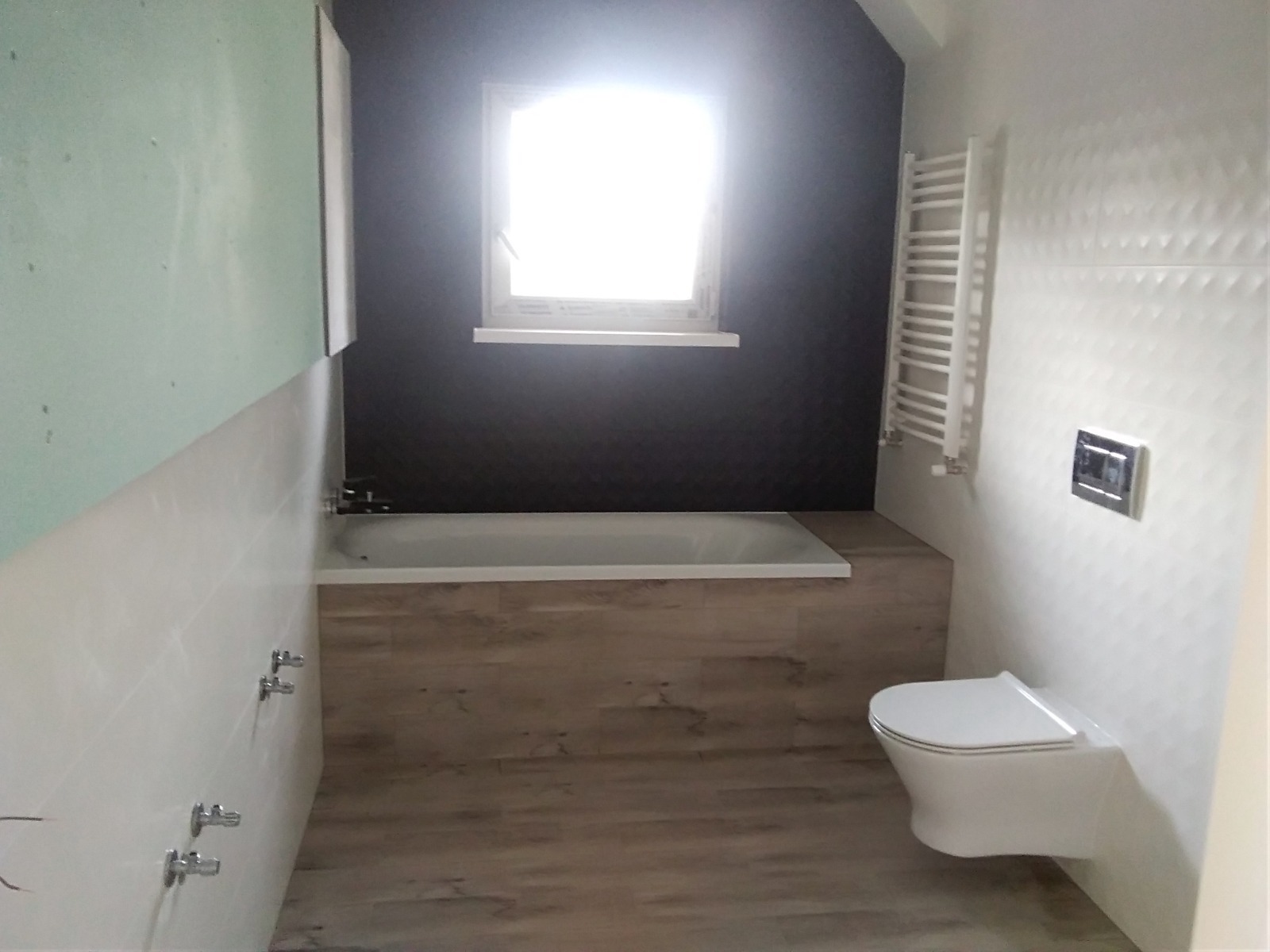 Wykończona łazienka z wanną obudowaną panelami imitującymi drewno, białą toaletą podwieszaną i grzejnikiem drabinkowym. Fragment ściany z płytą karton-gips.