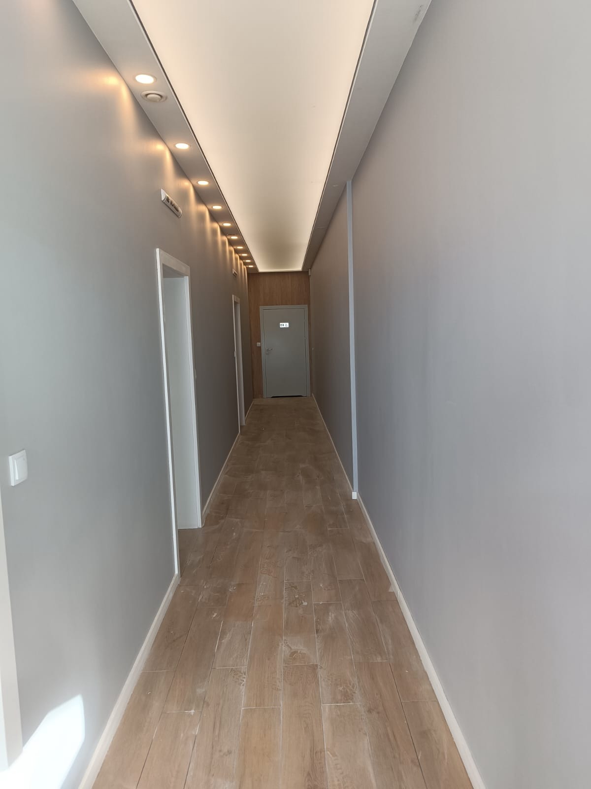 Korytarz po remoncie: szare ściany, drewnopodobna podłoga z płytek, oświetlenie sufitowe LED, drzwi wejściowe na końcu korytarza.