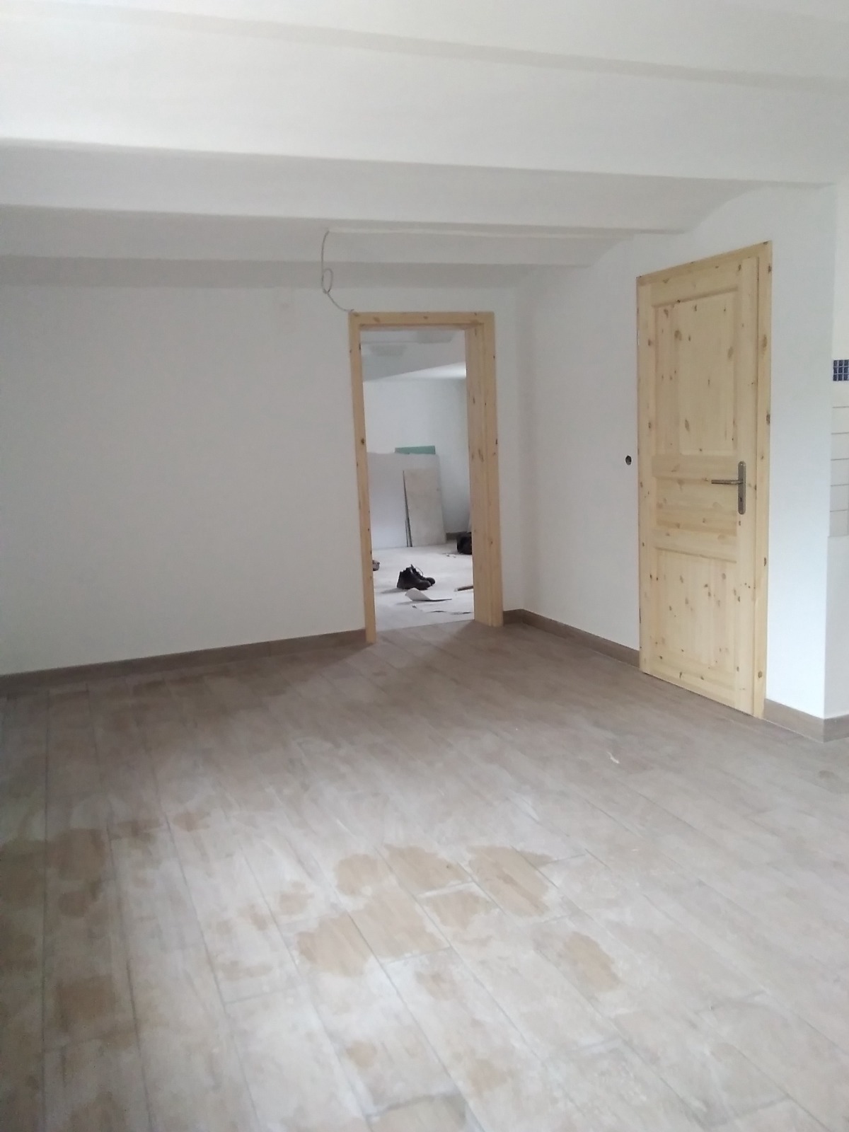 Jasne wnętrze po odświeżeniu: gładkie, białe ściany, drewniane drzwi i nowe panele podłogowe z widocznymi plamami. Widok na sąsiednie pomieszczenie przez otwarte drzwi.