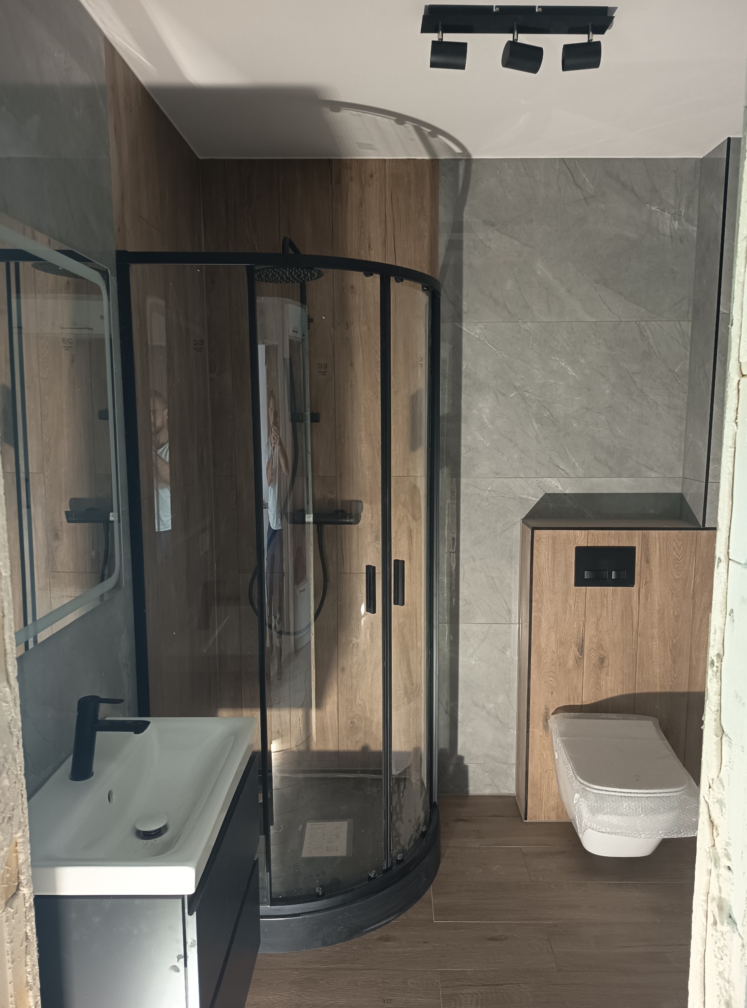 Wykończona łazienka z kabiną prysznicową, umywalką z szafką i toaletą w zabudowie. Płytki imitujące drewno i marmur. Oświetlenie punktowe.