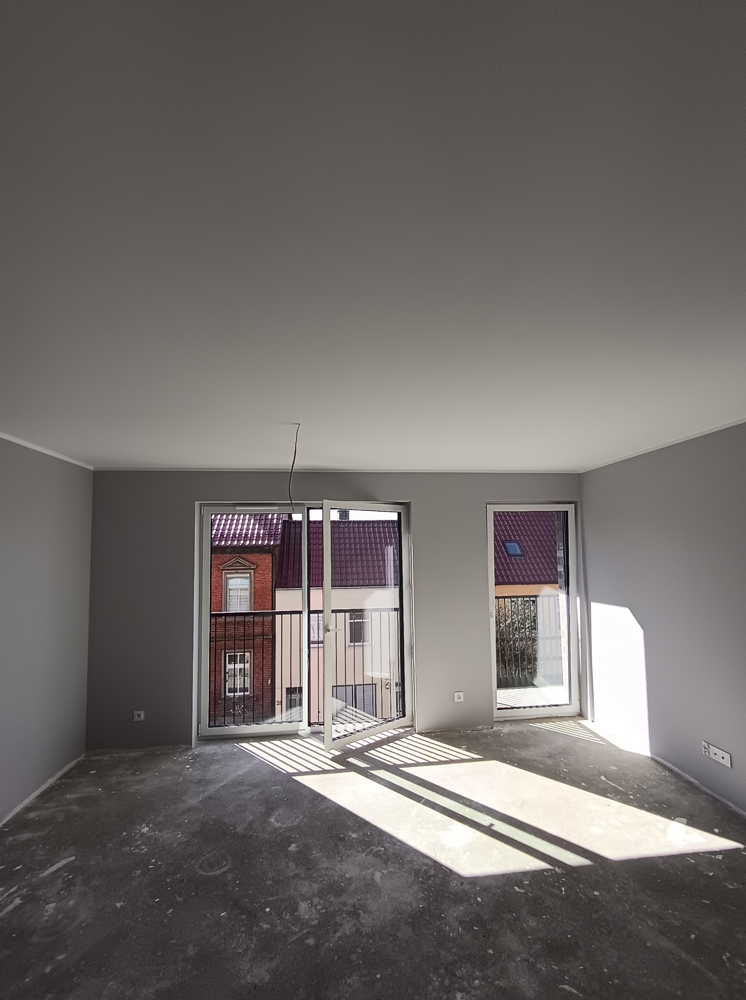 Pomalowane na szaro wnętrze pokoju z dużymi oknami balkonowymi i surową podłogą. Widoczne promienie słoneczne wpadające do środka. Świeżo wykończone ściany.