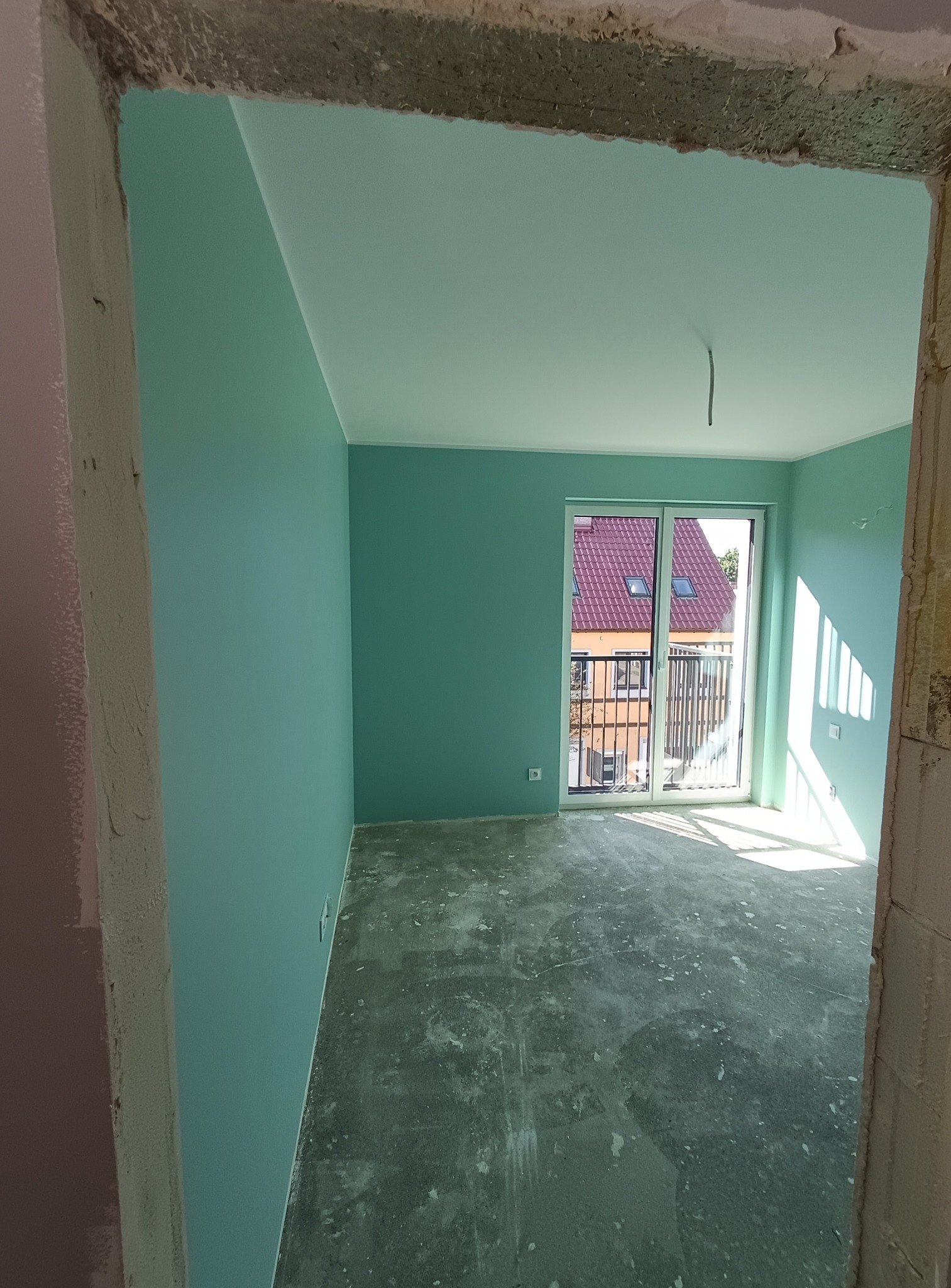 Pomieszczenie po malowaniu ścian na turkusowo, widoczne surowe wykończenie podłogi i ościeżnicy. W tle okno balkonowe z widokiem na budynek.