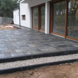 Nowo wyłożony taras z szarej kostki brukowej o nieregularnym kształcie, obrzeża z czarnego betonu i jasne kamyczki, widok na biały dom z dużymi przesuwnymi drzwiami tarasowymi.