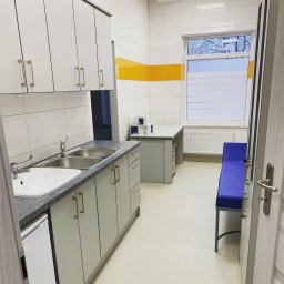 LuxeLike Sprzątanie Mycie Czyszczenie - Wnętrze gabinetu lekarskiego z umywalką, szafkami, stołem i niebieską kozetką, widoczne przez uchylone drzwi.