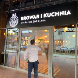 LuxeLike Sprzątanie Mycie Czyszczenie - Pracownik w Leżajsku myje okna wejściowe do restauracji 'Browar i Kuchnia', używając ściągaczki, widoczny napis 'Stara Ujeżdżalnia'.