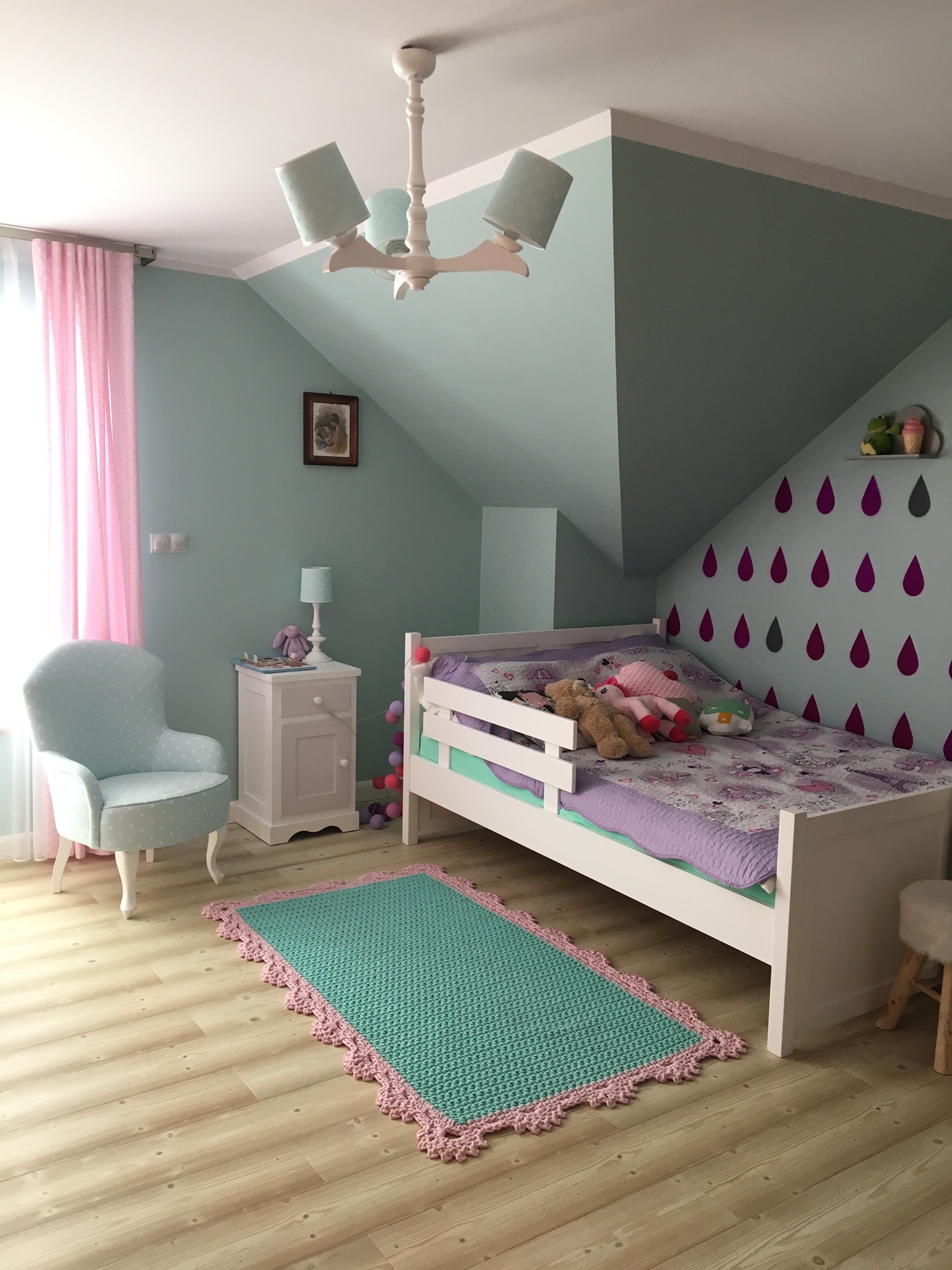Pokój dziecięcy w pastelowych kolorach z białym łóżkiem, turkusowym dywanem z różowym obramowaniem, ścianą w krople i drewnianą podłogą.