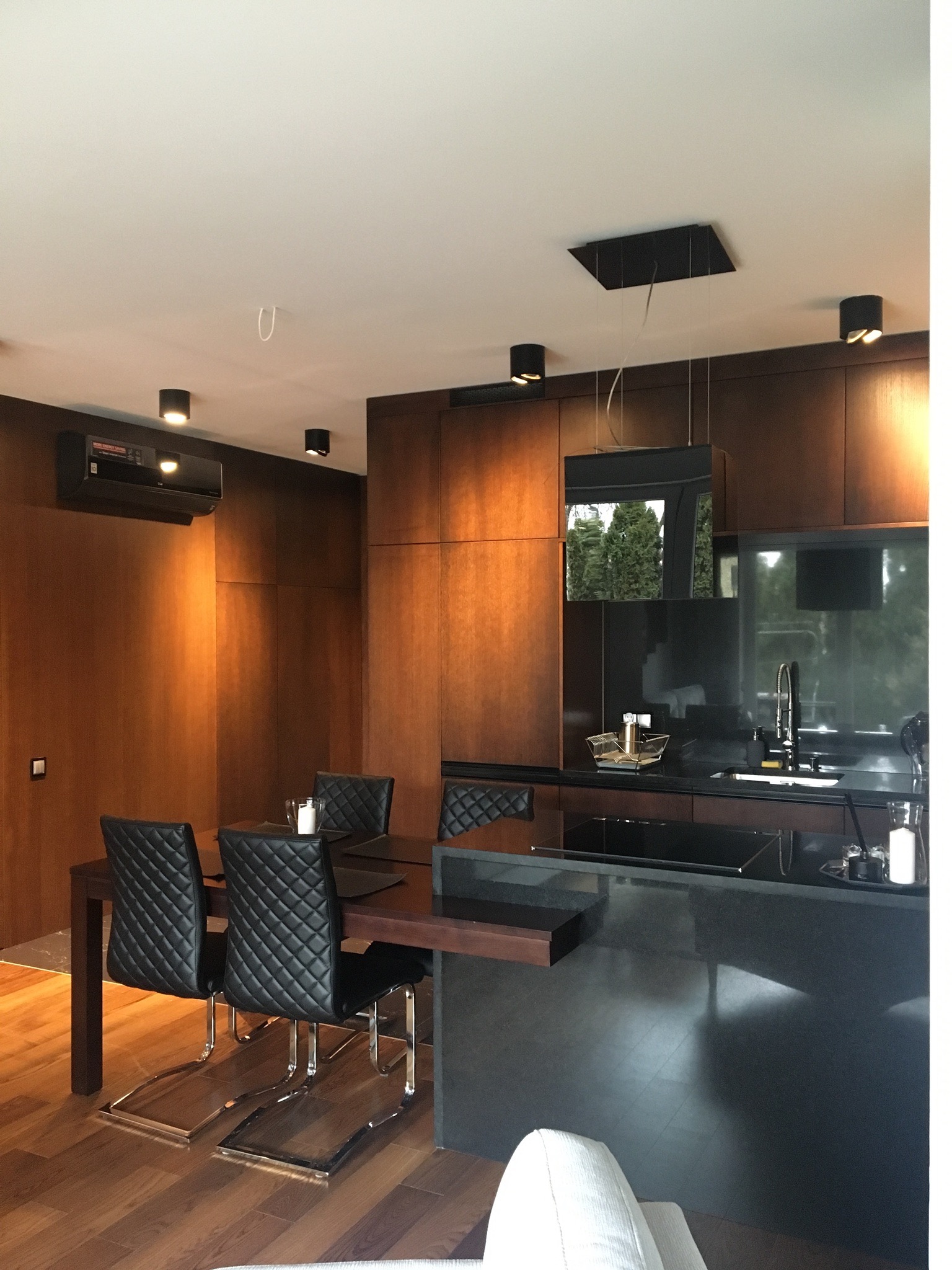 Nowoczesna kuchnia z ciemnego drewna i czarnymi akcentami, widoczne krzesła z pikowanym oparciem, minimalistyczne oświetlenie i drewniana podłoga.