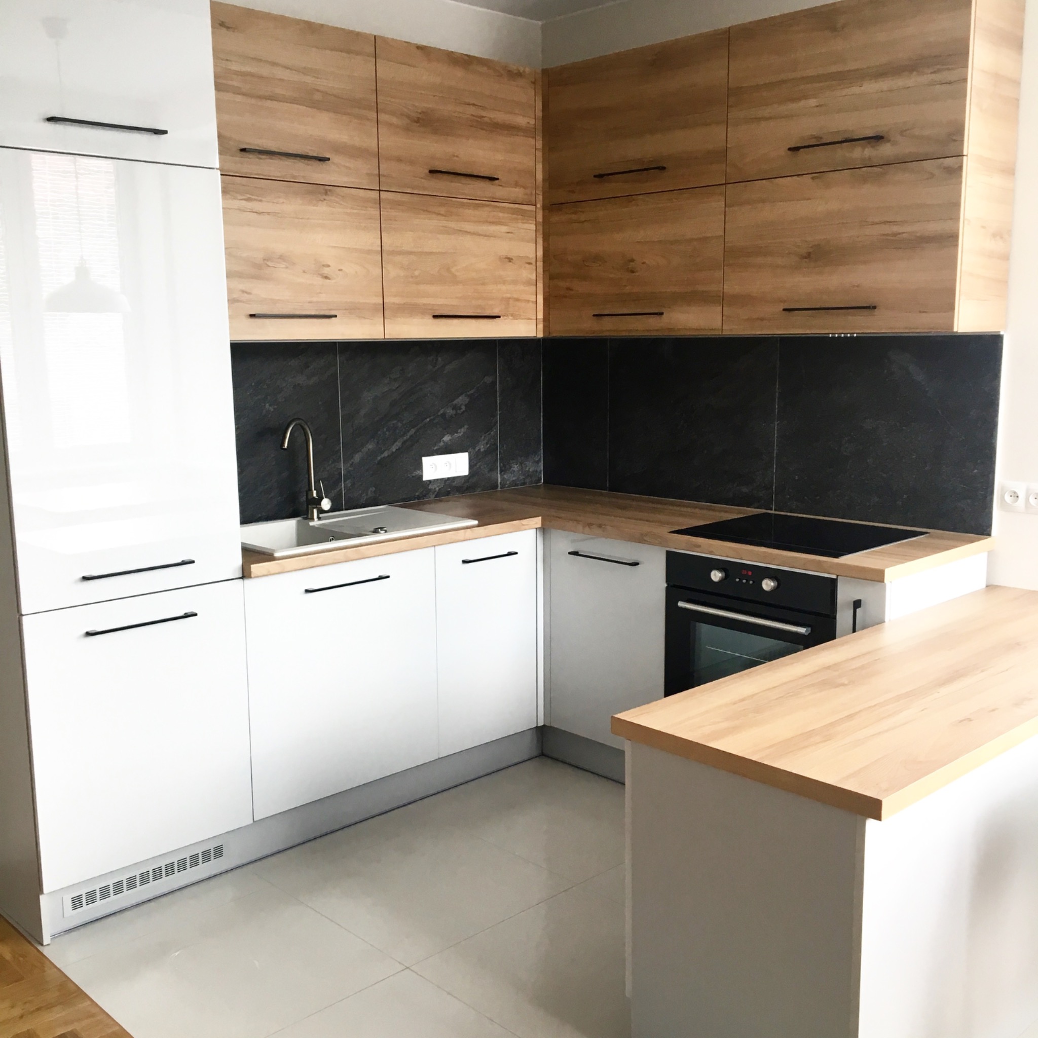 Nowoczesna kuchnia z białymi i drewnianymi szafkami, czarnym blatem i srebrną armaturą, oświetlona naturalnym światłem.