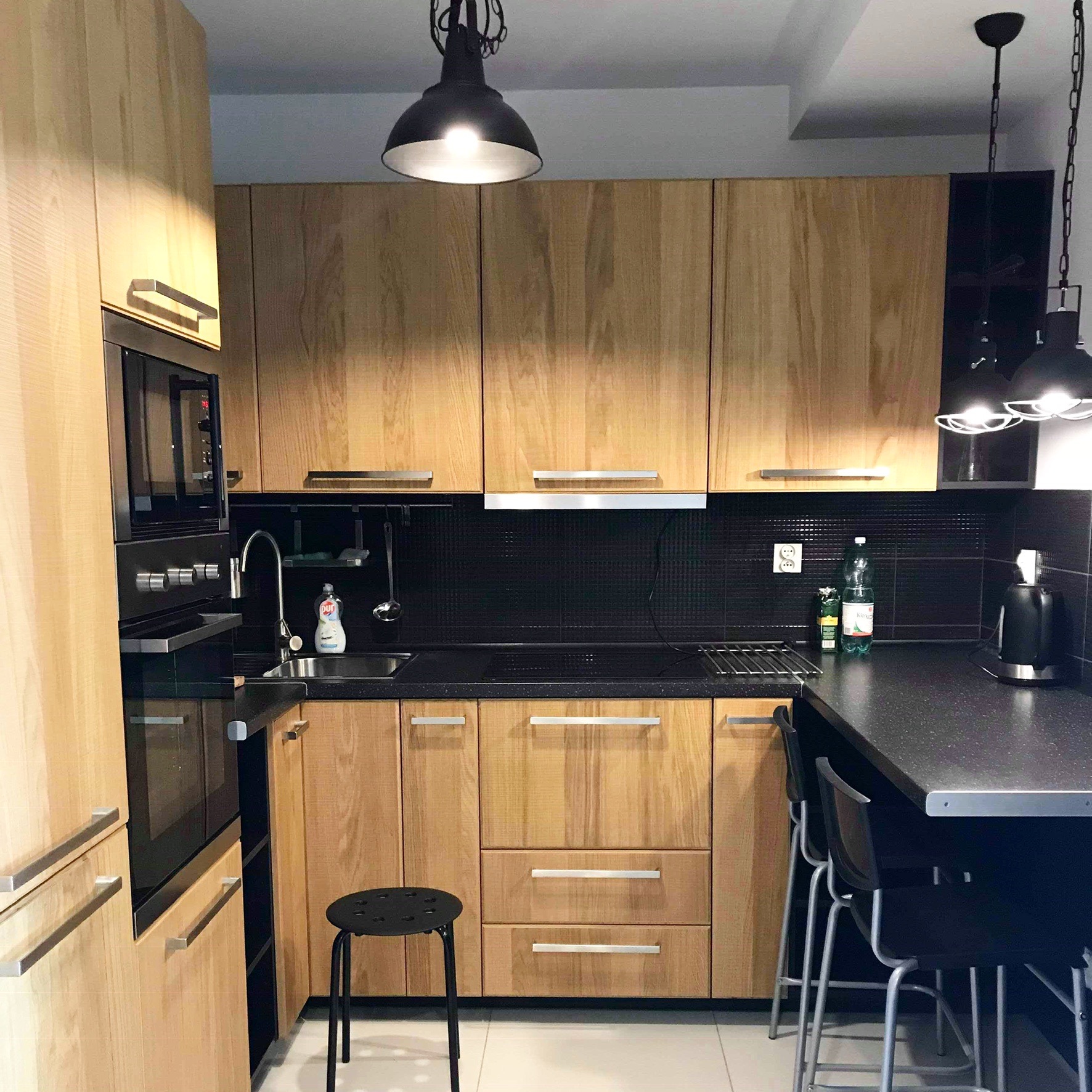 Jasna kuchnia z drewnianymi frontami, czarnym blatem i czarną mozaiką na ścianie, oświetlona wiszącymi lampami w stylu industrialnym.