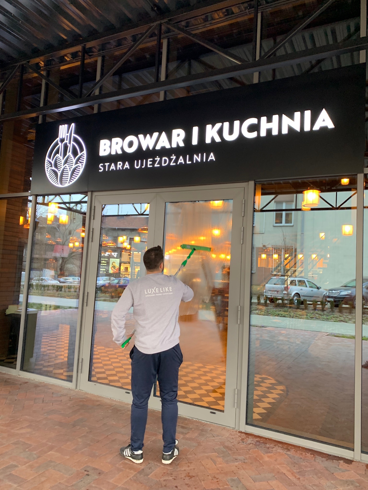 Pracownik w Leżajsku myje okna wejściowe do restauracji 'Browar i Kuchnia', używając ściągaczki, widoczny napis 'Stara Ujeżdżalnia'.