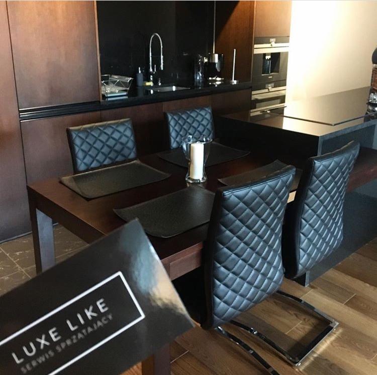 Elegancka jadalnia z ciemnymi meblami i czarnymi skórzanymi krzesłami z pikowaniem, na stole czarne podkładki i świeca, w tle widoczny aneks kuchenny, w rogu wizytówka firmy sprzątającej.