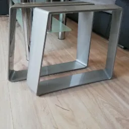 Dwie metalowe nogi do stołu o kwadratowym przekroju, z zaokrąglonymi narożnikami, ustawione na drewnianej podłodze, w tle fragment sofy i stolika.