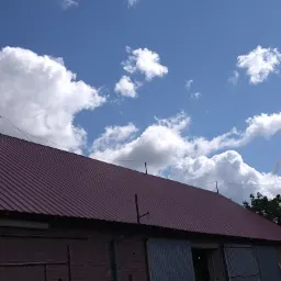 Czerwony blaszany dach budynku gospodarczego z ceglanymi ścianami, widziany pod kątem z wyraźnym błękitnym niebem i białymi obłokami w tle.