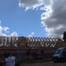 Dach-Mix - Budynek gospodarczy z drewnianą konstrukcją dachu w trakcie budowy, na tle błękitnego nieba z chmurami, obok zaparkowany czarny bus.