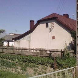 Dach-Mix - Dom jednorodzinny z czerwonym dachem widziany zza drewnianego płotu, z widocznym kominem i anteną na dachu.