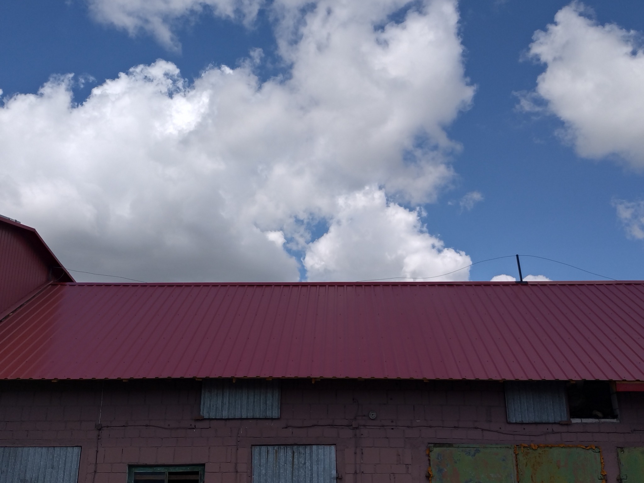 Czerwony blaszany dach na tle błękitnego nieba z białymi chmurami, widok na budynek z cegły z zamkniętymi okiennicami.