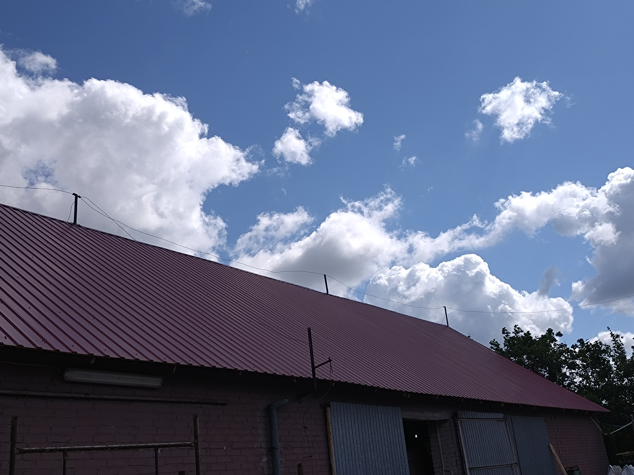 Czerwony blaszany dach budynku gospodarczego z ceglanymi ścianami, widziany pod kątem z wyraźnym błękitnym niebem i białymi obłokami w tle.