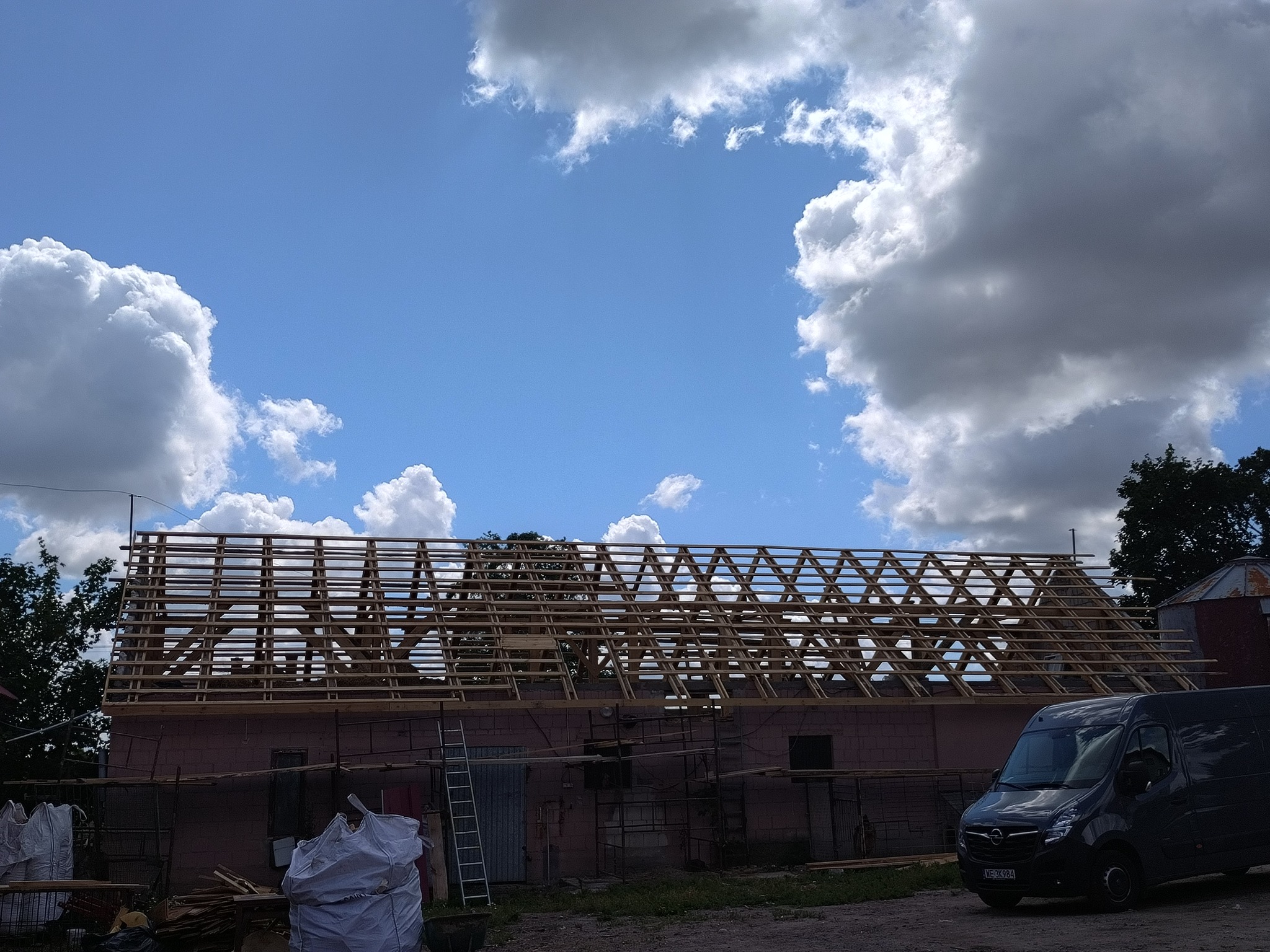 Konstrukcja więźby dachowej z drewnianych belek na budynku w trakcie budowy, widoczne worki z materiałami budowlanymi, drabina i ciemny samochód dostawczy na tle błękitnego nieba z chmurami.