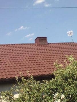 Czerwono-brązowe pokrycie dachowe z blachy trapezowej, widok z dołu, komin z brązową obudową, antena telewizyjna na dachu, błękitne niebo z chmurami w tle, częściowo zasłonięte przez gałęzie drzew.