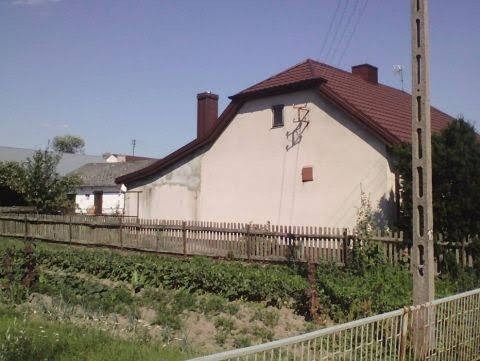 Dom jednorodzinny z czerwonym dachem widziany zza drewnianego płotu, z widocznym kominem i anteną na dachu.