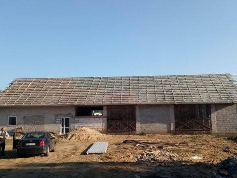 Budynek gospodarczy z częściowo zdjętym dachem, widoczna konstrukcja łatowania pod nowe pokrycie, na podwórzu zaparkowany samochód i składowane płyty.