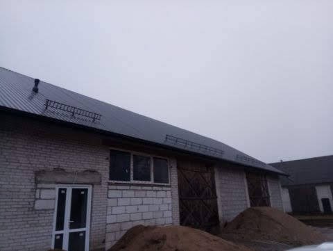 Metalowy dach na dużym budynku gospodarczym z cegły, widoczne zabezpieczenia przeciwśniegowe i komin, ujęcie z dołu w pochmurny dzień.