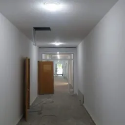 Korytarz w trakcie remontu, świeżo pomalowane białe ściany, widoczne otwory w suficie i kable na podłodze.