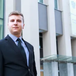 Młody mężczyzna w garniturze i krawacie, stojący na tle nowoczesnego budynku z elementami szkła i betonu.