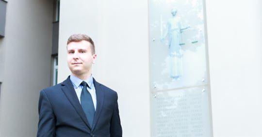 Młody mężczyzna w garniturze stoi na tle budynku z płaskorzeźbą Temidy, bogini sprawiedliwości.