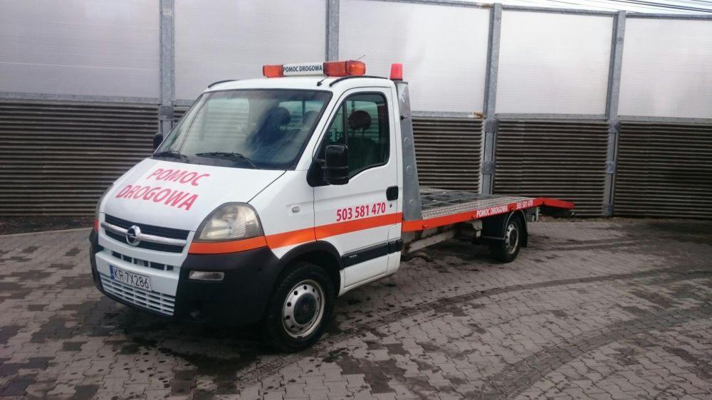 Biała laweta z pomarańczowymi akcentami i napisem 'Pomoc Drogowa' zaparkowana na kostce brukowej, z widocznym numerem telefonu na boku, na tle szarego, metalowego płotu.