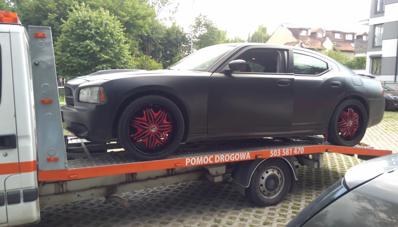 Matowy, czarny Dodge Charger z czerwonymi felgami transportowany na lawecie pomocy drogowej, widok z boku.