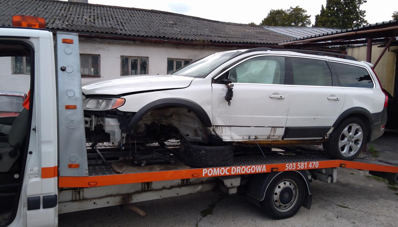 Uszkodzony biały samochód Volvo XC70 bez przedniego koła, transportowany na lawecie pomocy drogowej z widocznym numerem telefonu.