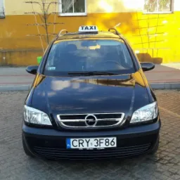 Czarna taksówka Opel Zafira z tablicą świetlną 'TAXI' na dachu, zaparkowana przed żółtym budynkiem z napisem 'Kwiaty' nad oknem.