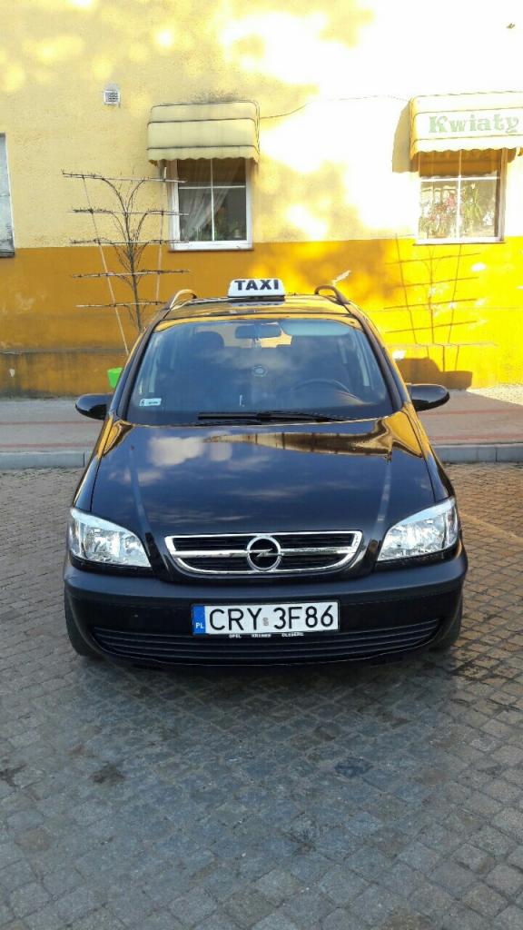 Czarna taksówka Opel Zafira z tablicą świetlną 'TAXI' na dachu, zaparkowana przed żółtym budynkiem z napisem 'Kwiaty' nad oknem.