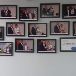 Ściana biura z ramkami prezentującymi zdjęcia osób odbierających certyfikaty lub nagrody, prawdopodobnie związane z dotacjami unijnymi, w tle jasna ściana.