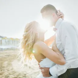 Para młoda w objęciach na plaży, silne światło słoneczne, panna młoda z blond lokami i długimi kolczykami, w tle budynki plażowe.