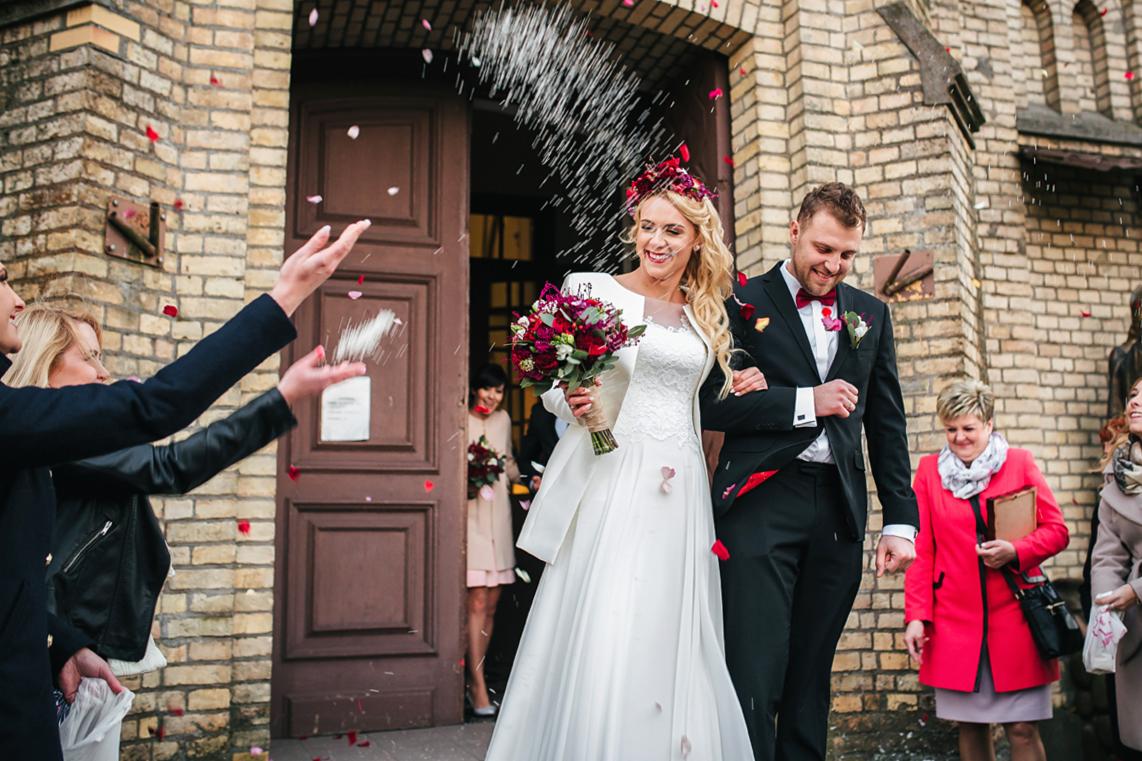 Para młoda opuszcza kościół, obsypywana płatkami róż, w tle ceglana fasada i uśmiechnięci goście weselni.